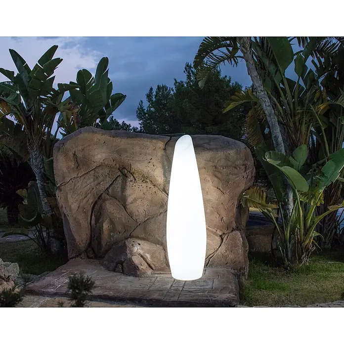 New Garden Lámpara de pie con batería Fredo 140 - 1,3 W, Blanco, Ø x Al: 38 x 139 cm, RGB - Lámpara de pie exterior luminosa blanca, alta y ovalada, situada junto a una roca en un jardín al anochecer.