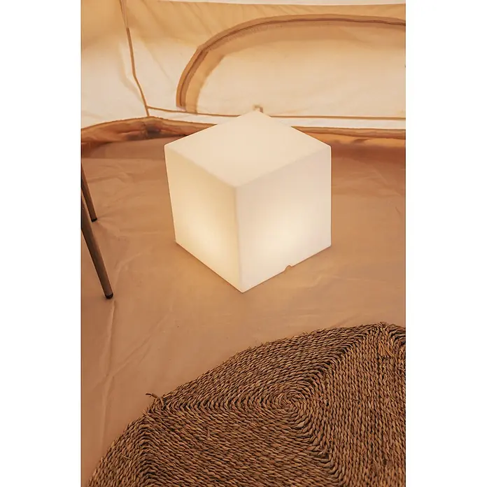 New Garden Lámpara solar Cuby 40 LED - 1 W, Blanco, Color de luz: RGBW, L x An x Al: 40 x 40 x 40 cm - Una lámpara de cubo blanca brillante se encuentra sobre un suelo de tienda marrón claro junto a una alfombra tejida, proporcionando luz ambiental.