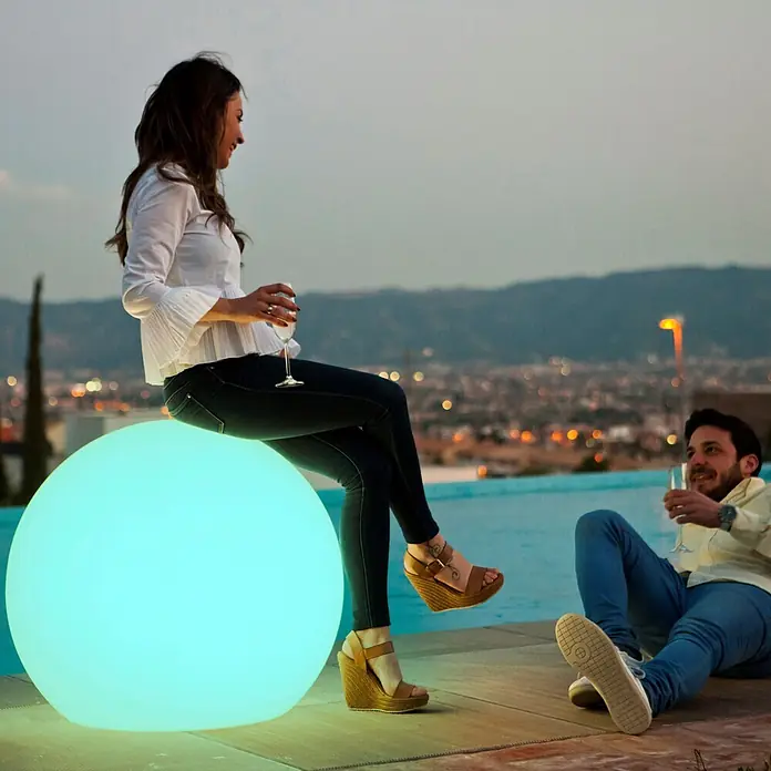 New Garden Lámpara decorativa redonda LED Buly 80 Solar - 5 W, Blanco, Diámetro: 57 cm - 78 cm, RGBW - Gran esfera luminosa azul claro que sirve de asiento exterior junto a una piscina al anochecer.