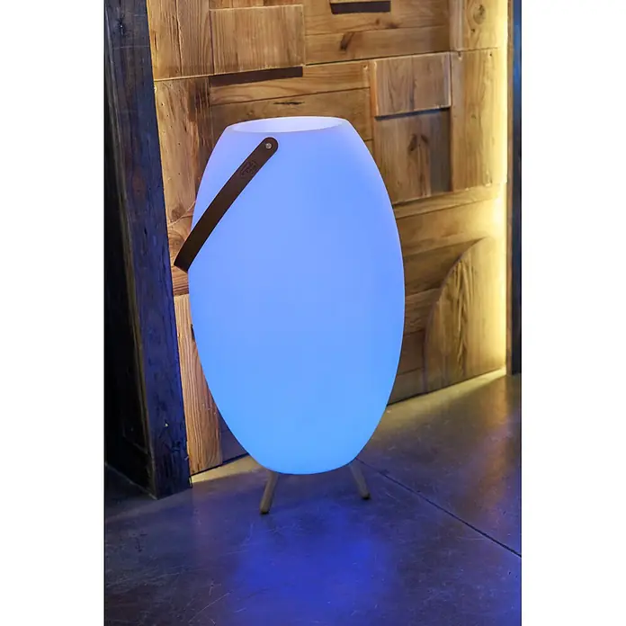 New Garden Lámpara para exterior con altavoz Apolo 70 - 6 W, Blanco, Ø x Al: 40 x 72,5 cm, RGBW, Altavoz - Lámpara ovalada portátil que brilla azul, con asa de cuero marrón y patas de madera, sobre una pared de madera rústica y suelo de hormigón.