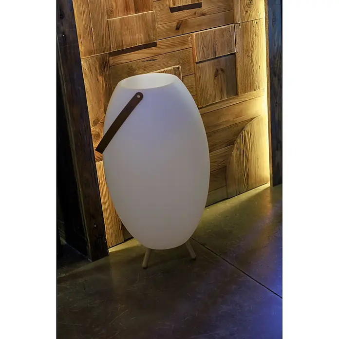 New Garden Lámpara para exterior con altavoz Apolo 70 - 6 W, Blanco, Ø x Al: 40 x 72,5 cm, RGBW, Altavoz - Una lámpara de pie ovalada blanca luminosa con asa marrón y patas de madera, sobre una pared de madera rústica.