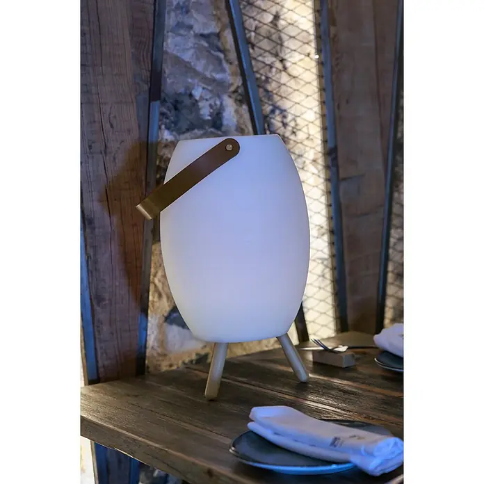 New Garden Lámpara para exterior con altavoz Apolo 45 - 6 W, Blanco, Ø x Al: 24 x 40,5 cm, RGBW, Altavoz - Lámpara de mesa ovalada blanca con patas de madera y asa de cuero marrón, iluminando una mesa de madera rústica.