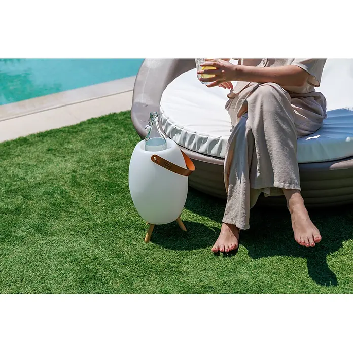 New Garden Lámpara para exterior con altavoz Apolo 45 - 6 W, Blanco, Ø x Al: 24 x 40,5 cm, RGBW, Altavoz - Nevera exterior blanca y ovalada con asa de cuero marrón y patas de madera, con una botella, junto a una persona relajándose en la piscina.