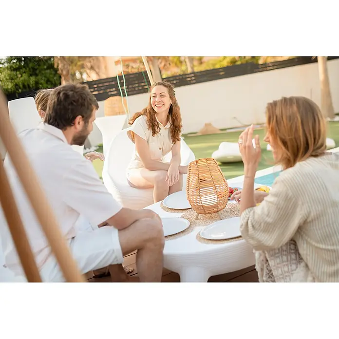 New Garden Bossa Lámpara para exterior a batería 30 - 9 W, Marrón, Blanco cálido, Ø x Al: 22,5 x 32,5 cm - Linterna de ratán natural tejida sobre una mesa, proporcionando luz ambiental para una reunión al aire libre.