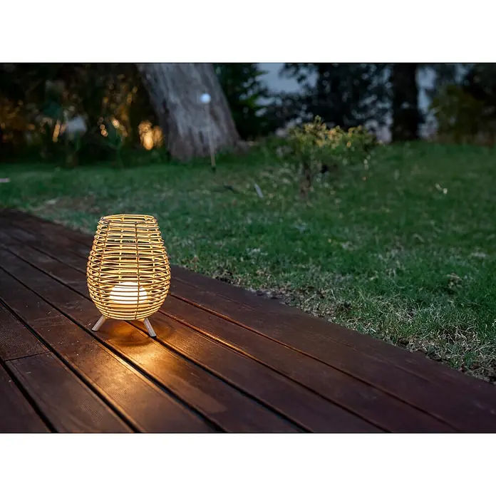 New Garden Bossa Lámpara para exterior a batería 30 - 9 W, Marrón, Blanco cálido, Ø x Al: 22,5 x 32,5 cm - Farol de ratán trenzado con luz cálida, iluminando una tarima de madera en un jardín nocturno.
