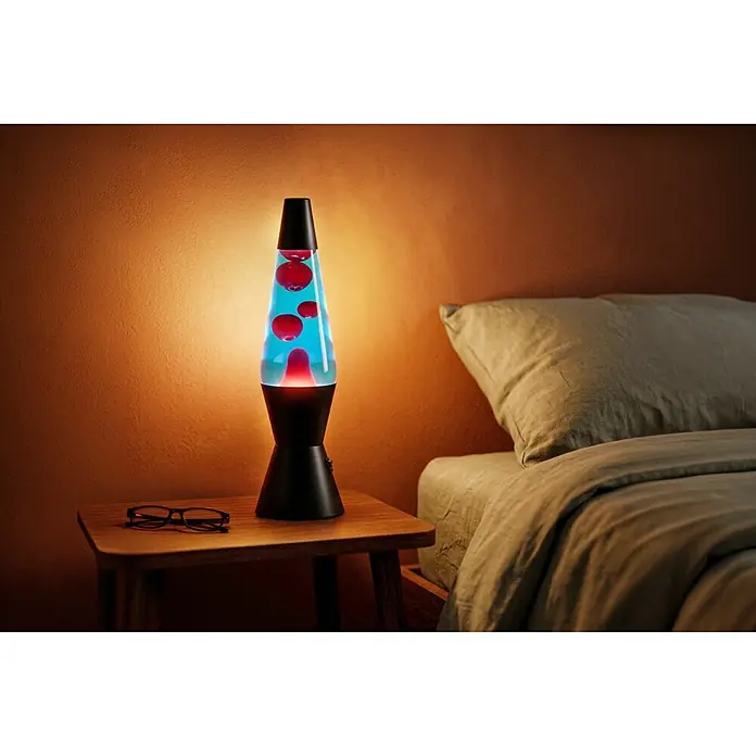 Eglo Lavalampe - 2,2 W, Ø x H: 105 x 360 mm, Farbe Gehäuse: Blau/ Rot - Eine schwarze Lavalampe mit roter 'Lava' in blauer Flüssigkeit steht auf einem Nachttisch in einem Schlafzimmer, neben einer Brille und einem Bett.