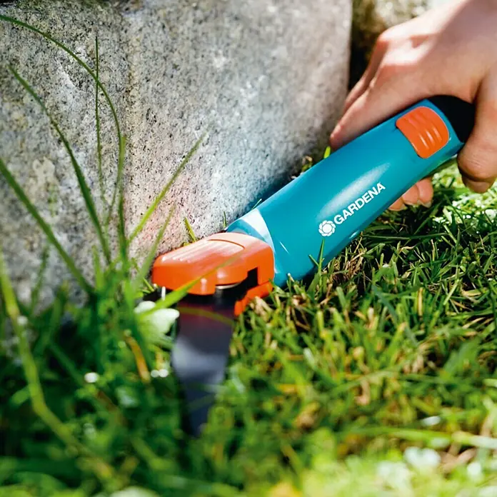 Gardena Comfort GrasschereMaterial Klinge: Geschliffene Schneide, Antihaftbeschichtet, Drehbar - Nahaufnahme einer Gardena Grasschere im Einsatz, um Rasen entlang einer Steinmauer zu trimmen.