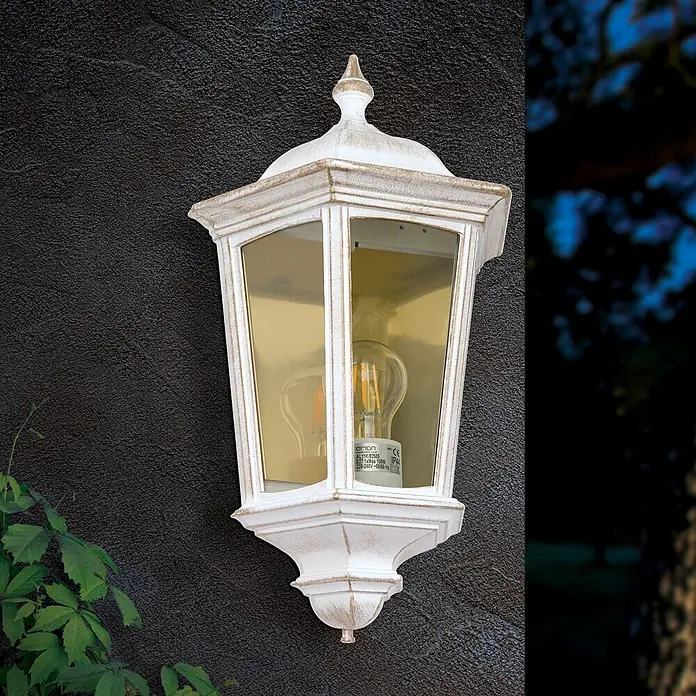Orion Leuchten Außenwandleuchte Puchberg - 60 W, Weiß, Klar, IP44 - Eine weiße, antik wirkende Außenwandleuchte mit Glühfadenlampe an einer dunklen strukturierten Wand neben grünen Blättern.