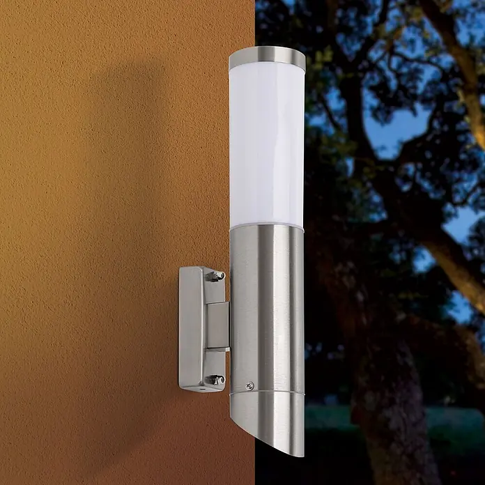 Orion Leuchten Außenwandleuchte Pleno - 13 W, Silber, Opal, IP44 - Wandleuchte aus gebürstetem Edelstahl mit weißem zylindrischem Diffusor, an einer strukturierten braunen Wand montiert, beleuchtet eine nächtliche Außenszene.