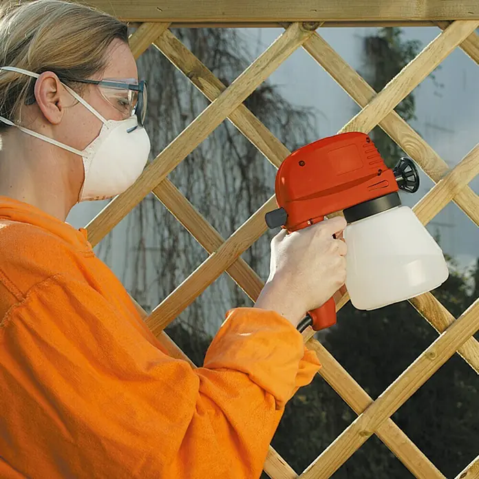 Lehnartz Elektrische Spritzpistole Easy-Sprayer PLUS - 45 W - Frau mit Schutzmaske streicht einen Holzzaun mit einem orangefarbenen Farbspritzgerät.