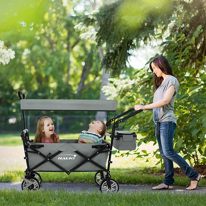Hauki Bollerwagen Handwagen Transportwagen faltbar klappbar mit Bremse Dach und Tasche - L x B x H: 196 x 56 x 99 cm - Grauer Hauki Bollerwagen mit Verdeck, zwei lachende Kinder transportierend, von einer Frau im Freien geschoben.