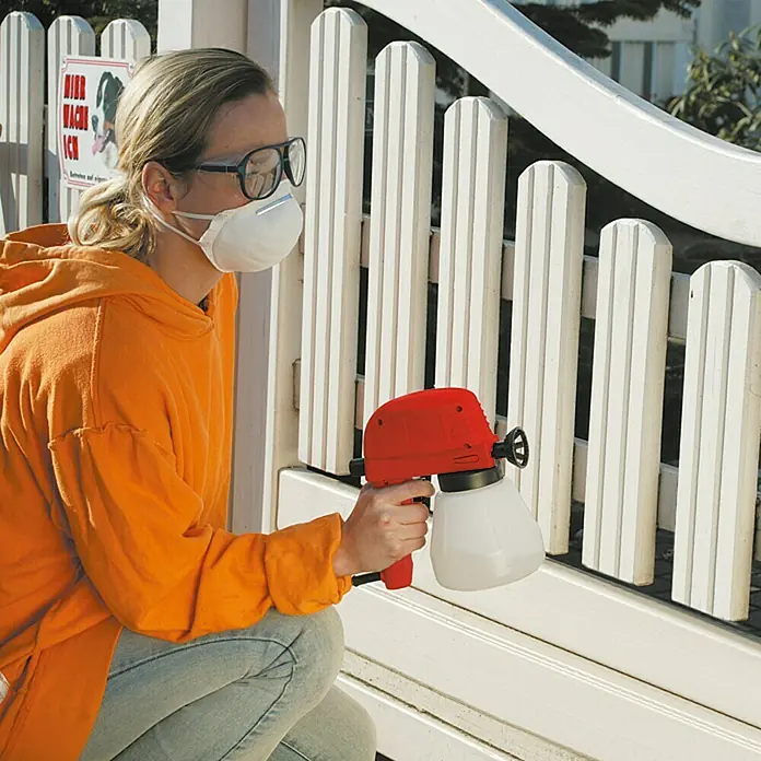 Lehnartz Elektrische Spritzpistole Easy-Sprayer PLUS - 45 W - Eine Person benutzt eine rote Farbspritzpistole mit weißem Behälter, um einen weißen Holzzaun zu streichen. Sie trägt eine Maske und eine Brille.