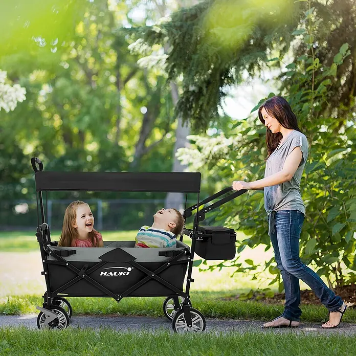 Hauki Bollerwagen Handwagen Transportwagen faltbar klappbar mit Bremse Dach und Tasche - L x B x H: 196 x 56 x 99 cm - Schwarzer und grauer Faltwagen mit Verdeck, der zwei Kinder befördert und von einer Frau im Freien geschoben wird.