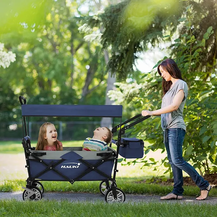 Hauki Bollerwagen Handwagen Transportwagen faltbar klappbar mit Bremse Dach und Tasche - L x B x H: 196 x 56 x 99 cm - Eine Frau schiebt einen blau-grauen Bollerwagen mit zwei fröhlichen Kindern auf einem Parkweg.