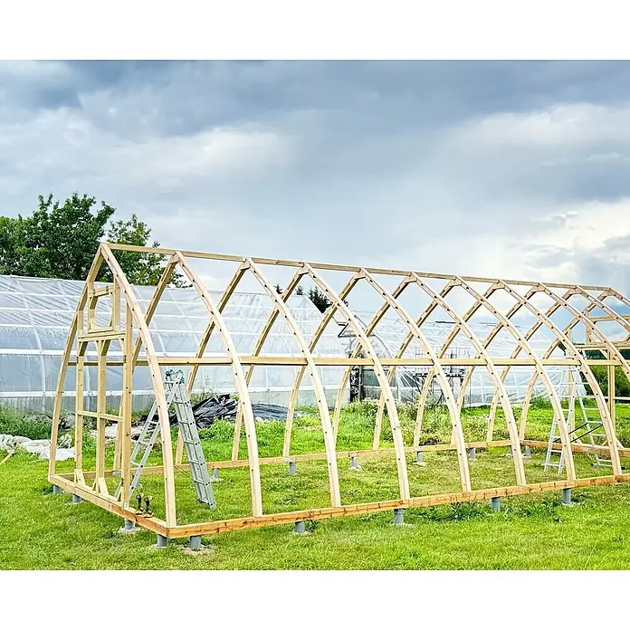 Garten Dream Gewächshaus Arched Wood - Außenmaß inkl. Dachüberstand (B x T): 500 x 1.000 cm, Material Seitenwand: Kunststoff, 4 mm, Braun - Großes Holzgewächshausgestell im Bau auf einer Wiese. Zeigt die beträchtliche Größe und das Naturholz.