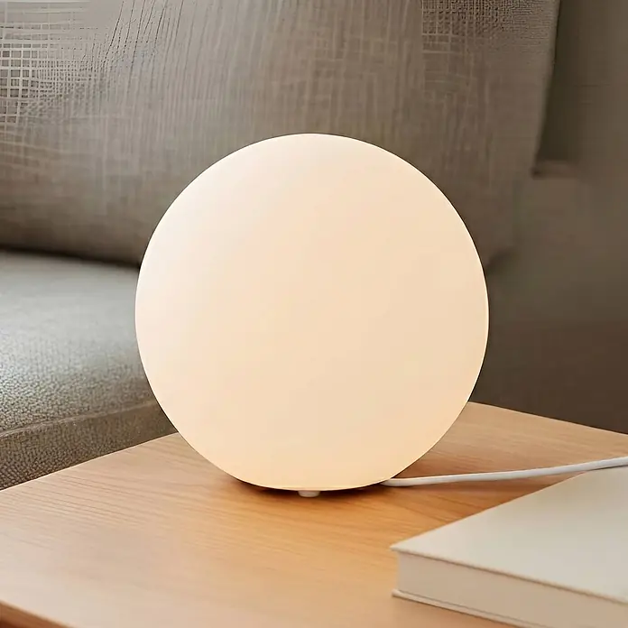 Lindby Tischleuchte Marike - weiß / opal, E27 - Eine beleuchtete weiße Kugellampe steht auf einem hellen Holztisch neben einem Buch, mit einem beigen Sofa im Hintergrund.