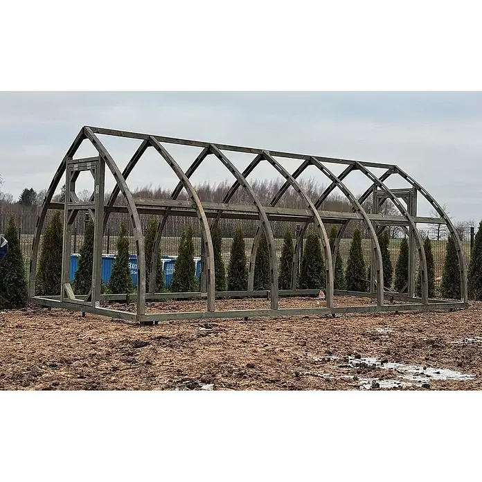 Garten Dream Gewächshaus Arched Wood - Außenmaß inkl. Dachüberstand (B x T): 300 x 400 cm, Material Seitenwand: Kunststoff, 4 mm, Braun - Graues Holzbogengewächshausgestell auf schlammigem Boden, Bäume im Hintergrund.