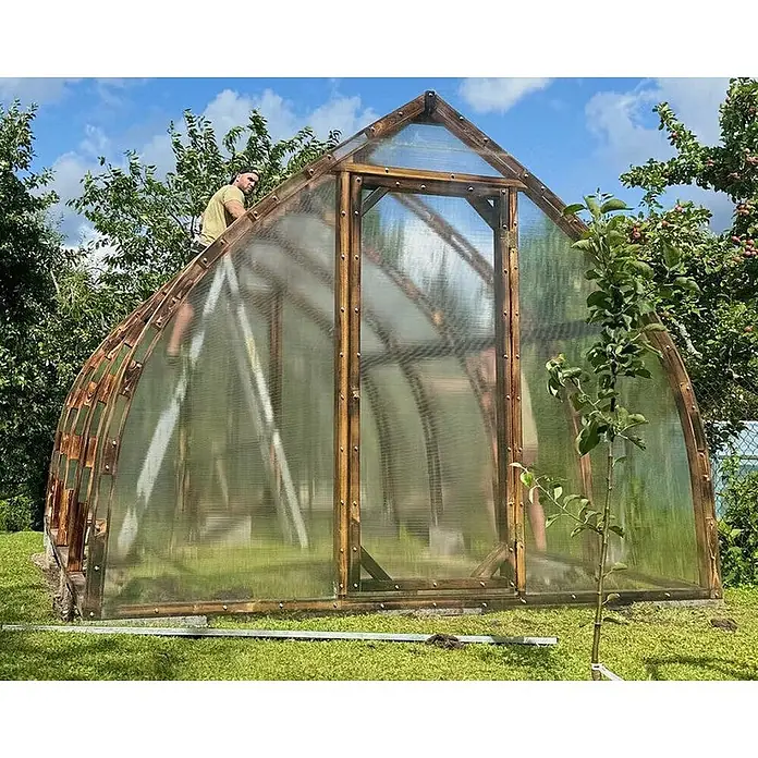 Garten Dream Gewächshaus Arched Wood - Außenmaß inkl. Dachüberstand (B x T): 300 x 400 cm, Material Seitenwand: Kunststoff, 4 mm, Braun - Großes gewölbtes Gewächshaus mit dunklem Holzrahmen und transparenten Paneelen, mit einer Person bei der Montage oder Wartung.