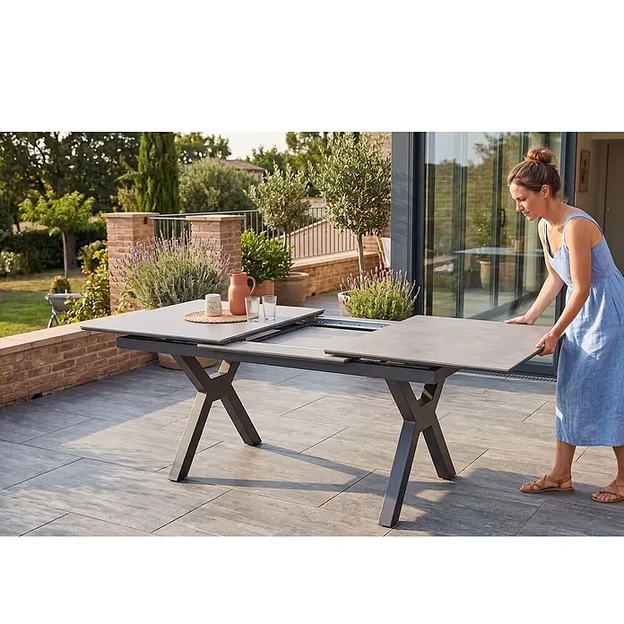 Bene Living Gartentisch Granada - L x B x H: 210 x 90 x 76 cm, Aluminium Gestell mit gesinnerter Tischplatte aus Keramik, Rechteckig, Anthrazit - Frau erweitert einen grauen ausziehbaren Esstisch für den Außenbereich auf einer Terrasse.