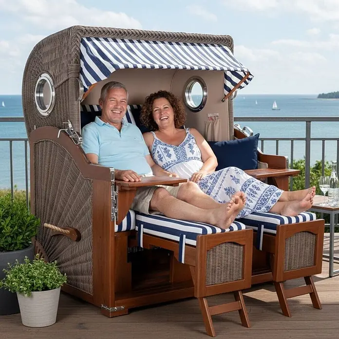 Mr. Deko Strandstuhl Strandkorb Düne Grande Bullauge Mahagoni PE Grey Dessin 518 - Teakholz. Polyrattan, Rückenlehne verstellbar, Blau, B x T x H: 142 x 95 x 170 cm - Ein Strandkorb aus Rattan und Holz mit blau-weiß gestreiften Polstern, in dem ein Paar am Meer entspannt.