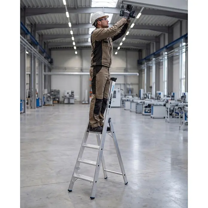 Krause Stehleiter XL 1x6 - Anzahl Tritte: 6 Stk., Arbeitshöhe: 230 cm, Belastbarkeit: 225 kg, Aluminium - Mann auf einer silbernen Stehleiter, der in einer Industriehalle über Kopf arbeitet.