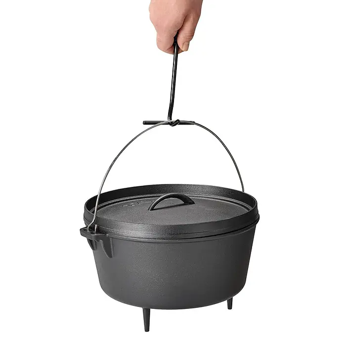 TAINO Dutch Oven - Ø x H: 30 x 22,5 cm, L x B x H: 33,5 x 35,5 x 22,5 cm, Gusseisen - Hand hält einen schwarzen Gusseisen-Dutch-Oven mit Füßen und Deckel mittels eines speziellen Henkelhebers.