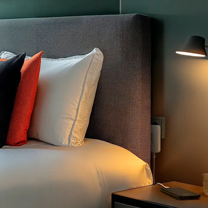 Hama Cargador rápido 00201976 - Blanco - Cabecero tapizado gris oscuro con almohadas blancas y naranjas en una cama, iluminado por una lámpara de noche.