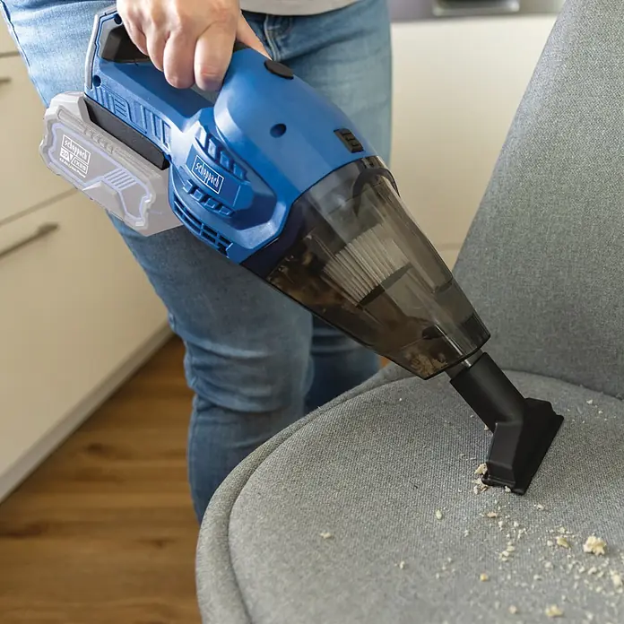 Scheppach IXES 20V Akku-Handstaubsauger C-HVC150-X - 20 V, Ohne Akku, Behältervolumen: 0,56 l - Blauer und schwarzer Scheppach Akku-Handstaubsauger reinigt Krümel von einem grauen Stuhl.