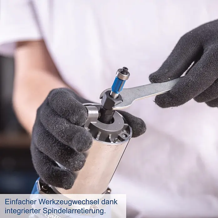 Scheppach IXES 20V Akku-Oberfräse BC-EHR24-X - 20 V, Ohne Akku, 10.000 U/min - 30.000 U/min, 8 mm - Hände in schwarzen Handschuhen wechseln einen blauen Fräser mit einem Schraubenschlüssel an einem silbernen Elektrowerkzeug, was den einfachen Werkzeugwechsel mit Spindelarretierung zeigt.