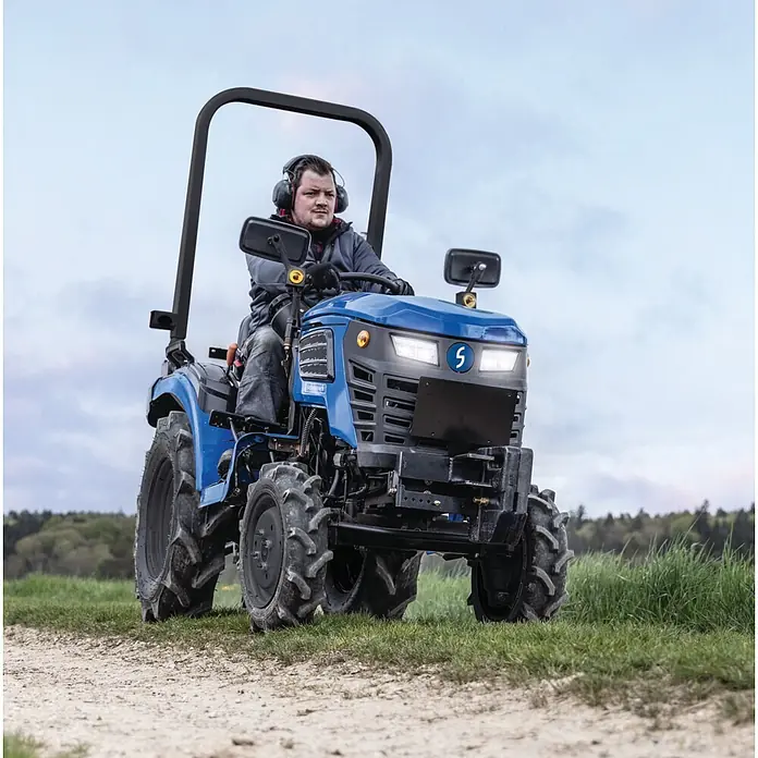Scheppach Kleintraktor MT25 - Hubraum: 1.132 cm³, Tankvolumen: 10 l - Ein Mann mit Gehörschutz fährt einen blauen Kompakttraktor mit schwarzen Reifen auf einem Feldweg neben grünem Gras.