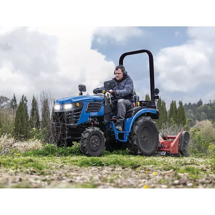 Scheppach Kleintraktor MT25 - Hubraum: 1.132 cm³, Tankvolumen: 10 l - Ein Mann bedient einen blauen Scheppach Traktor mit rotem Anbaugerät auf einem grasbewachsenen Feld. Der kompakte Traktor ist im Einsatz zu sehen.