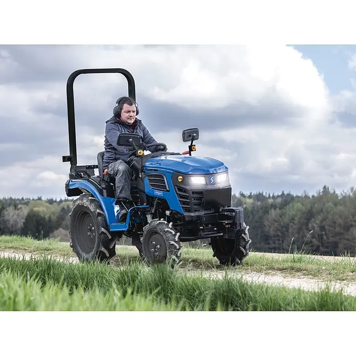 Scheppach Kleintraktor MT25 - Hubraum: 1.132 cm³, Tankvolumen: 10 l - Eine Person mit Gehörschutz fährt einen blauen Scheppach Kompakttraktor auf einem Feldweg.