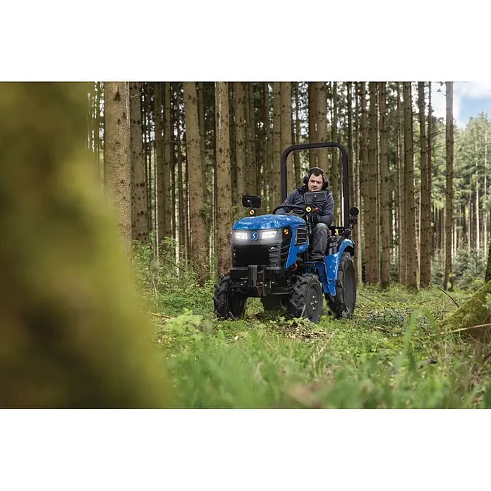 Scheppach Kleintraktor MT25 - Hubraum: 1.132 cm³, Tankvolumen: 10 l - Person fährt blauen Kompakttraktor mit hellen Scheinwerfern durch einen Wald.