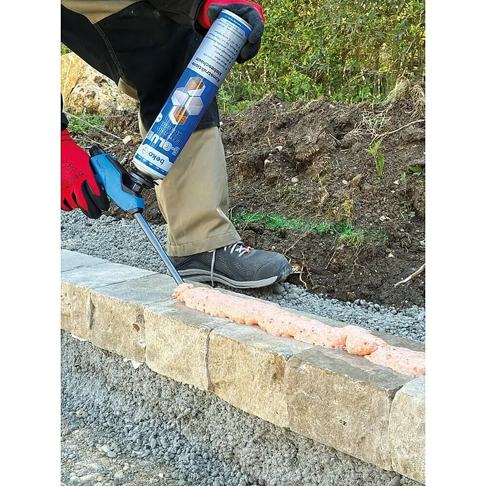 Beko Klebeschaum E-Glue - 810 ml - Person trägt orangefarbenen beko E-GLUE Konstruktions-Klebeschaum mit einer Pistole auf Steinblöcke zum Mauerkleben auf.