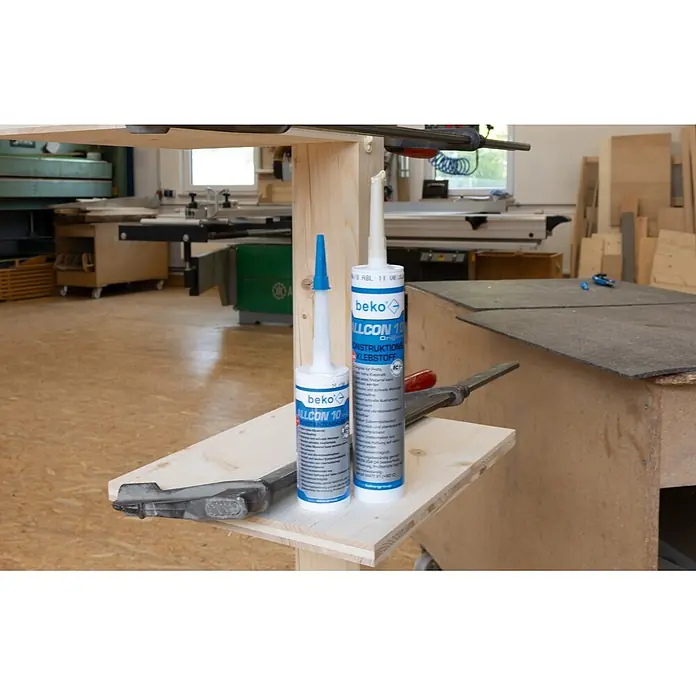 Beko Konstruktionskleber Allcon 10 - 150 ml, Beige - Zwei Tuben Beko Allcon 10 Konstruktionsklebstoff, eine mit blauer und eine mit weißer Kappe, auf einer Holzoberfläche in einer Werkstatt.