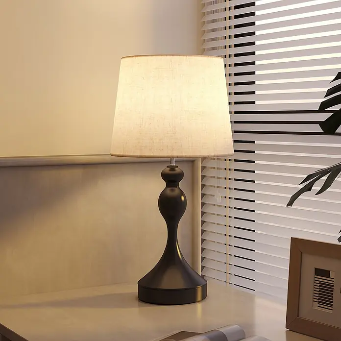 Homcom Tischleuchte - 40 W, L x B x H: 28 x 28 x 54 cm, Schwarz, Cremeweiß, E27 - Beleuchtete Tischlampe mit dunklem, verziertem Sockel und hellem Stoffschirm auf heller Fläche, die ein warmes Licht im Raum wirft.