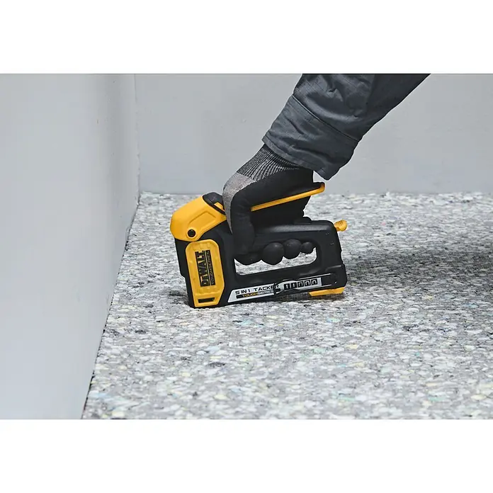DeWalt Grapadora manual DWHT70510 - Profundidad de grapado: 6 mm - 14 mm - Una mano enguantada presiona una grapadora DeWalt 5-en-1 amarilla y negra sobre el acolchado de la alfombra junto a una pared.