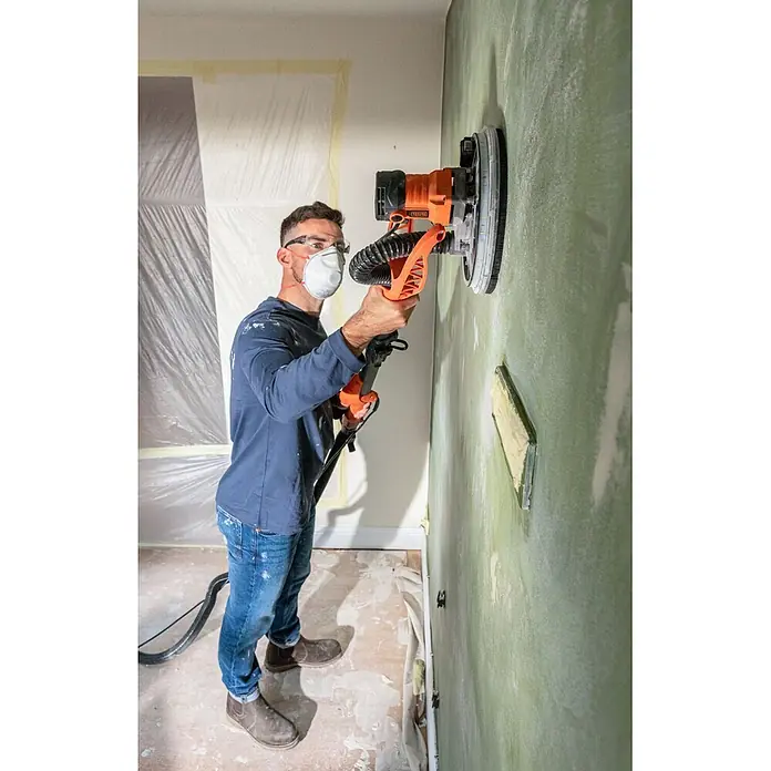 Black+Decker Lijadora de pared y techo BEDW700 - 710 W, Diámetro disco de lija: 225 mm - Un hombre con equipo de protección utiliza una lijadora de pared naranja y negra para alisar una pared verde.