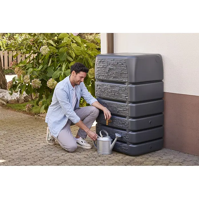 4rain Regentonne Mauertank Slim Stone - 510 l, Graphite grey - Mann füllt Gießkanne aus dunkelgrauem Regenwassertank mit Steinoptik für die Gartenbewässerung.