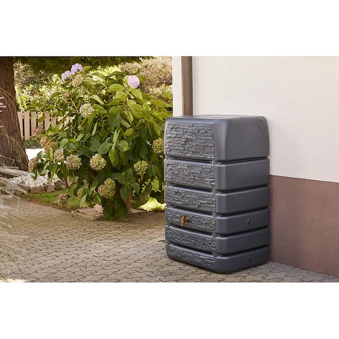 4rain Regentonne Mauertank Slim Stone - 510 l, Graphite grey - Dunkelgraue Regentonne mit Steintextur und Messinghahn, im Freien an einer Hauswand zur Wassersammlung platziert.