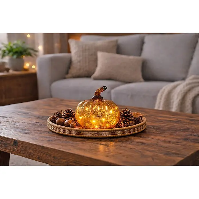 LED-Dekofigur Dekokürbis - L x B x H: 12 x 12 x 11,5 cm - Ein bernsteinfarbenes, beleuchtetes Glaskürbis-Centerpiece auf einem Weidentablett mit Tannenzapfen und Eicheln auf einem Holztisch im Wohnzimmer.