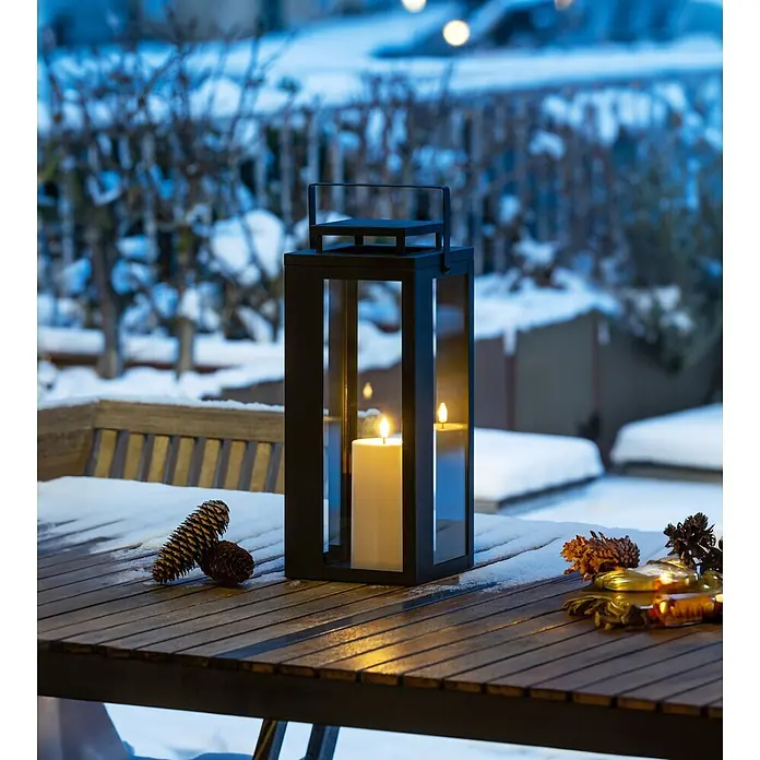 LED-Laterne Lucido - 16,5 x 16,5 x 40,5 cm, Schwarz - Schwarze Metalllaterne mit brennender Kerze auf einem verschneiten Holztisch im Freien, mit Tannenzapfen dekoriert.