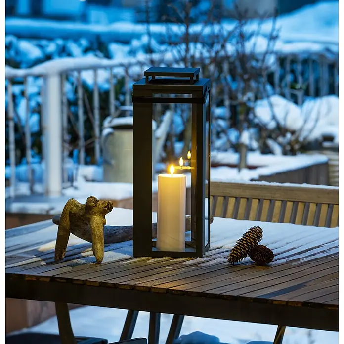 LED-Laterne Lucido - 18 x 18 x 48,5 cm, Schwarz - Schwarze Metalllaterne mit brennender Kerze auf einem schneebedeckten Holztisch, schafft eine gemütliche Winterstimmung.