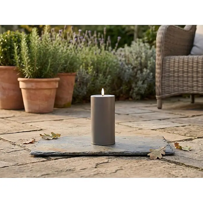 LED-Kerze Niumi - Ø x H: 8 x 15 cm, Greige, Metall - Eine brennende graue Kerze auf einer Schieferplatte, die das Ambiente einer Terrasse im Freien bereichert.