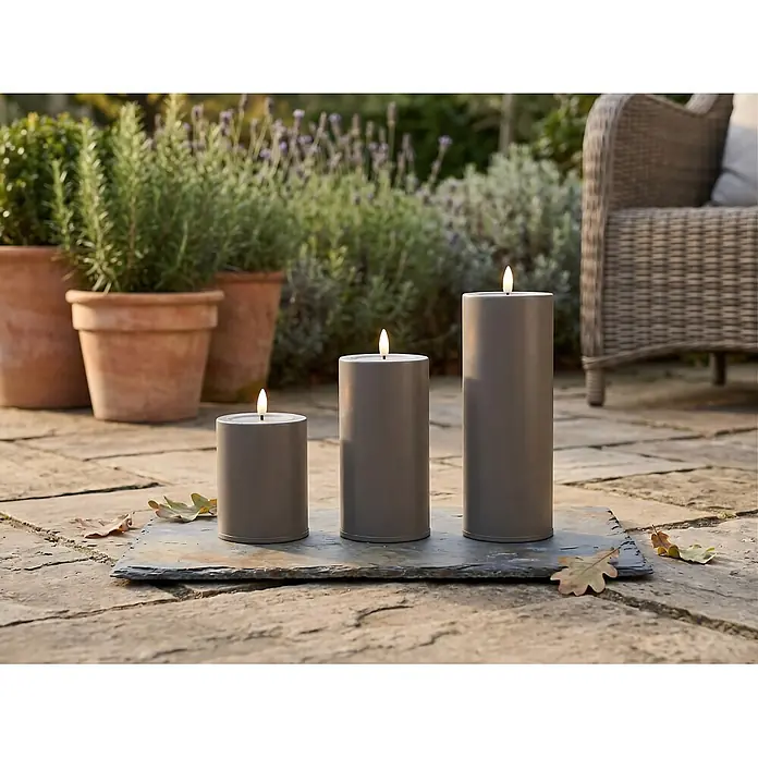 LED-Kerze Niumi - Ø x H: 8 x 10 cm, Greige, Metall - Drei graue zylindrische Outdoor-Kerzen auf einer Schieferplatte, die eine Terrasse beleuchten.