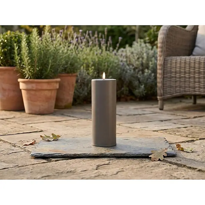 LED-Kerze Niumi - Ø x H: 8 x 20 cm, Greige, Metall - Eine hohe, graue zylindrische Outdoor-Kerze brennt auf einer Schieferplatte, auf einer Terrasse mit Topfpflanzen und einem Korbstuhl im Hintergrund.