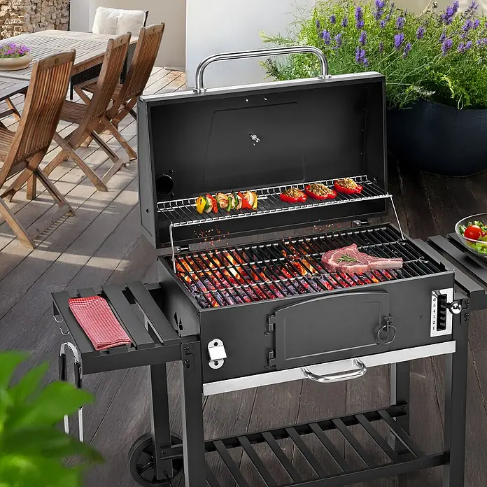 TAINO Holzkohlegrill HERO XXL Set - Schwarz - Schwarzer Holzkohlegrill aus Metall mit kochendem Essen, Beistelltisch und Rädern, auf einer Außenterrasse.
