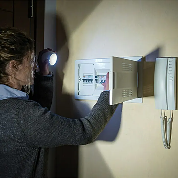 LED-Bewegungsmelder Vigilamp - Weiß, Außen - Eine Frau benutzt eine Taschenlampe, um einen Stromkasten an einer Wand zu inspizieren. Die weiße Sicherungsbox ist geöffnet und Telefon hängt daneben.
