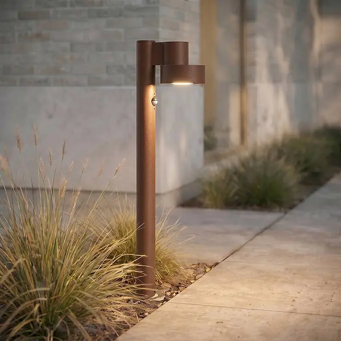 Lucande LED Wegeleuchte in braun, mit Bewegungsmelder, Gartenlampe -  - Rostfarbene Wegeleuchte mit integriertem LED und Bewegungssensor, die einen gepflasterten Weg und Gartenpflanzen beleuchtet.