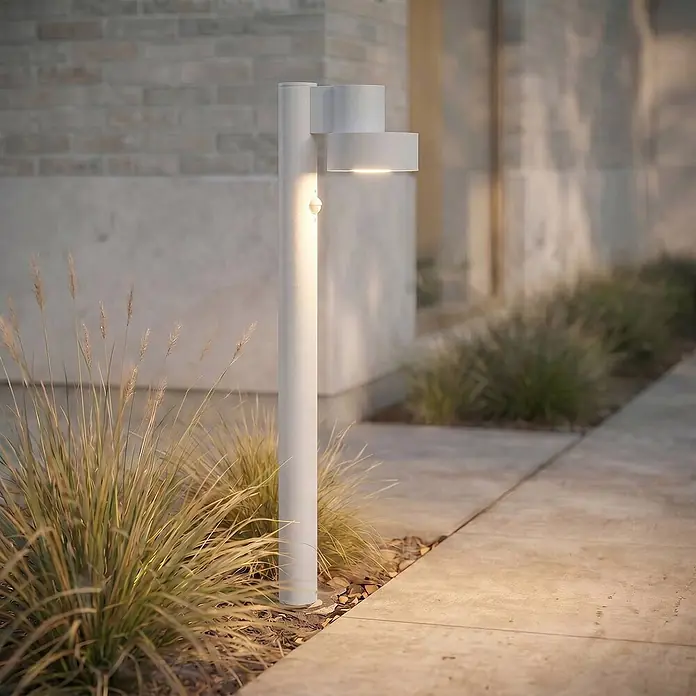 Lucande LED Wegeleuchte in hellgrau, mit Bewegungsmelder, Gartenlampe -  - Moderne hellgraue Pollerleuchte mit integriertem Bewegungssensor, beleuchtet einen Betonweg neben Ziergräsern.